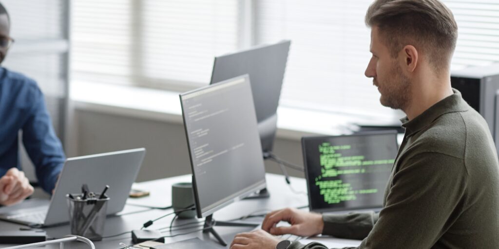 Man working at a desk with dual monitors displaying code in a modern office—illustrating cybersecurity work and the need for SME cyber insurance.