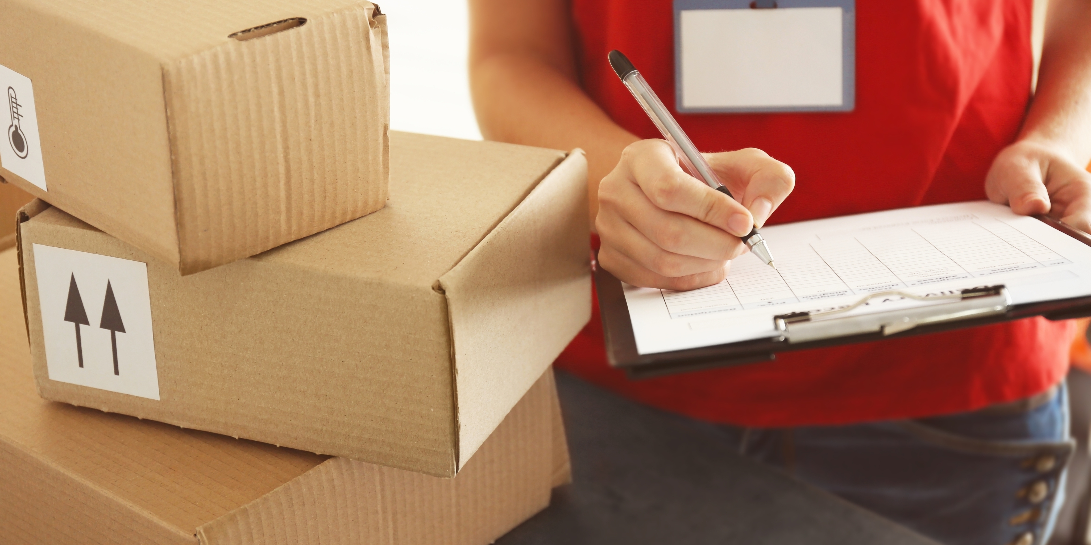 Courier in a red shirt checks a delivery manifest on a clipboard beside stacked cardboard boxes with handling labels.